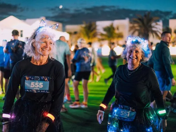 Two participants in glowing headbands and LED wristbands smile on the lawn at dusk, palm trees behind