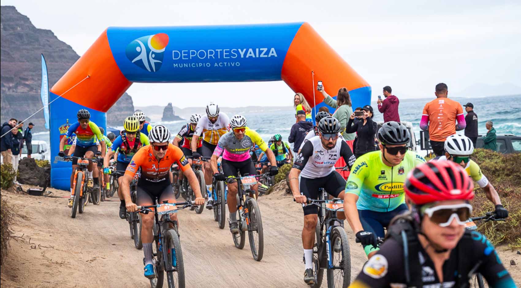 Mountain bike mass start under the Deportes Yaiza arch, on a sandy track beside the sea and the cliffs